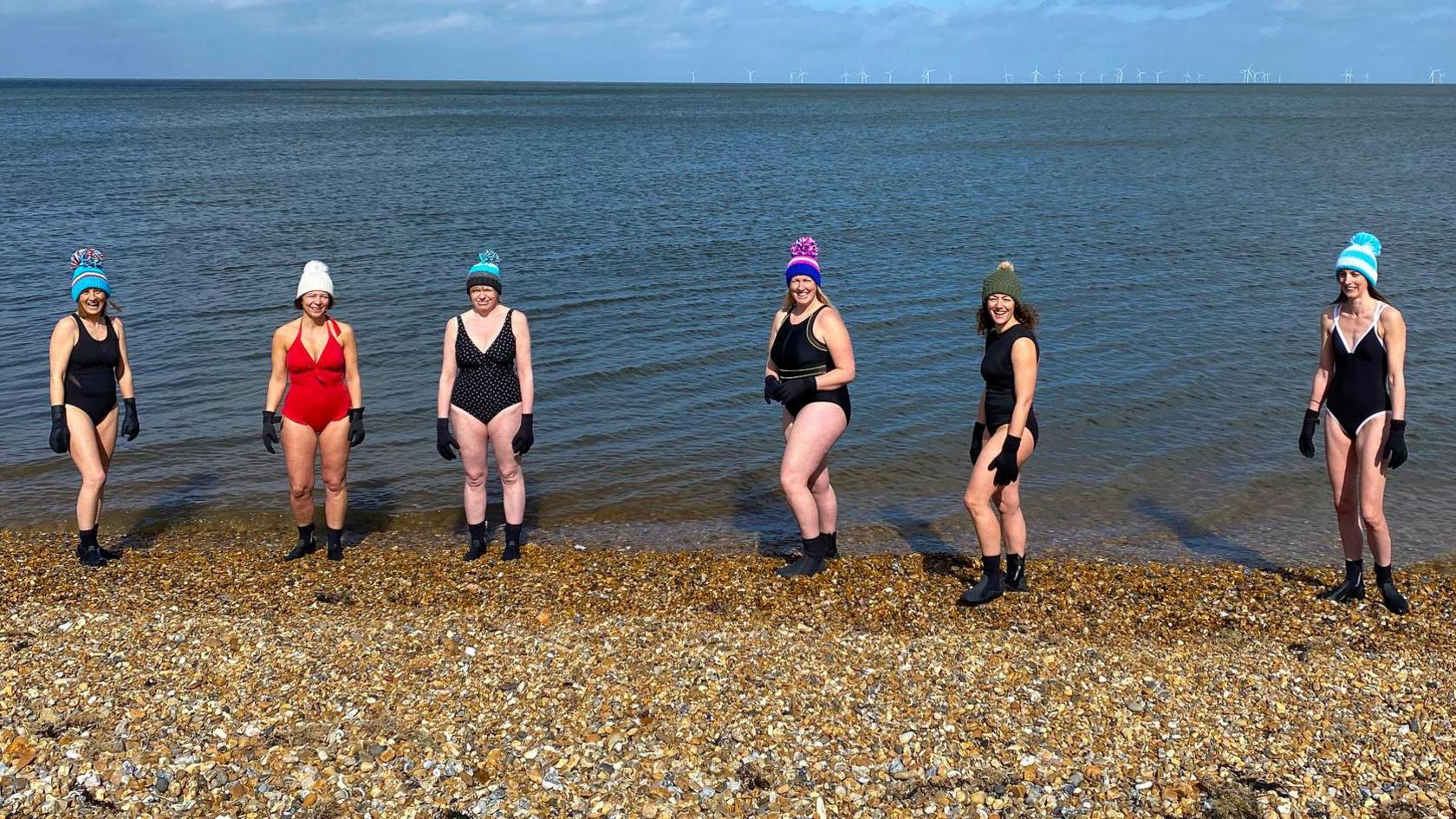 Six mature women standing on a pebbly beach by the sea. They are wearing swimming costumes, woollen hats, black gloves and ankle-high water shoes.