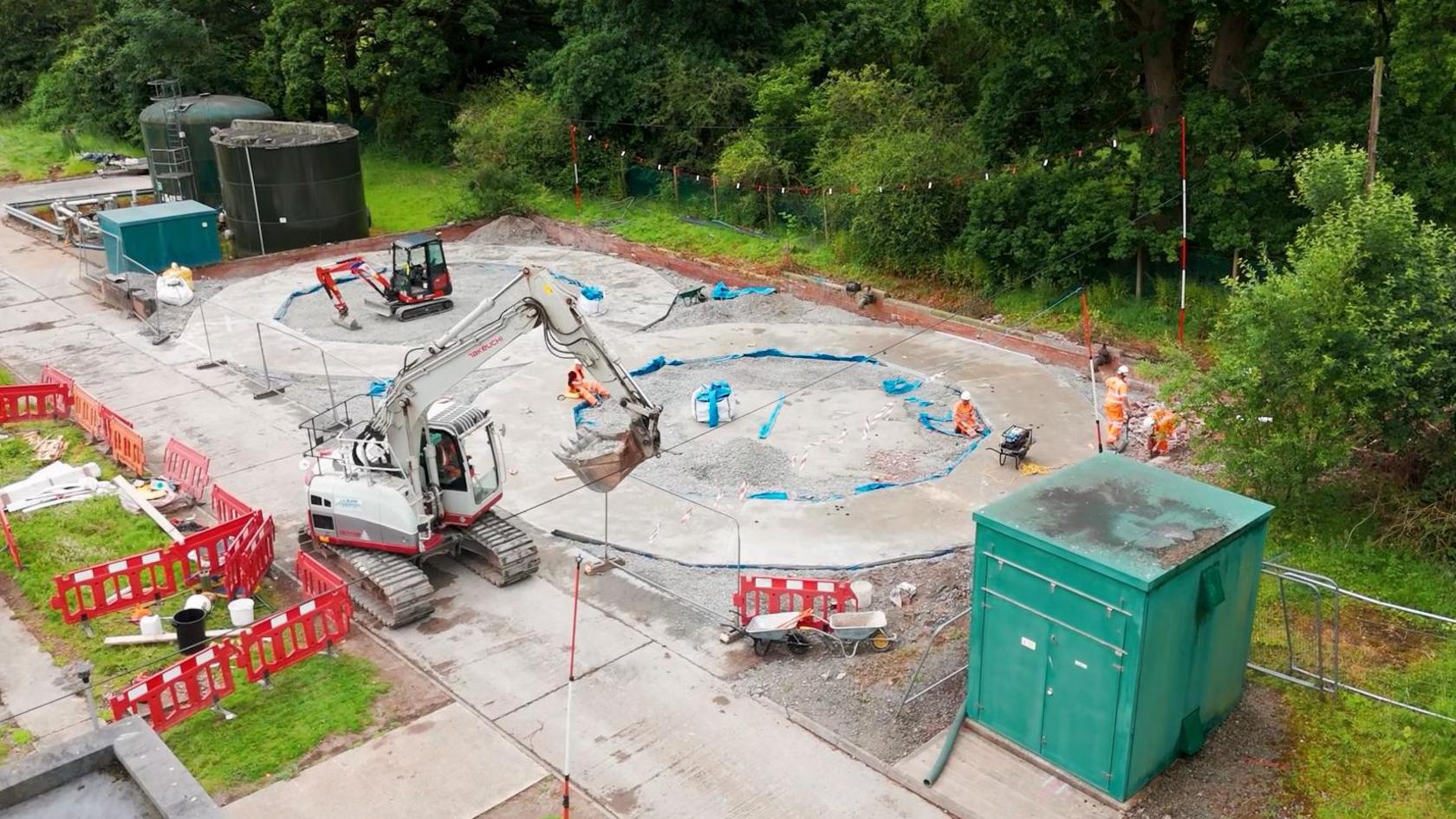 Photo of a storm tank being constructed. The foundations are being laid for 2 circular structures. There are 2 diggers in the shot, wheelbarrows and safety barriers.    