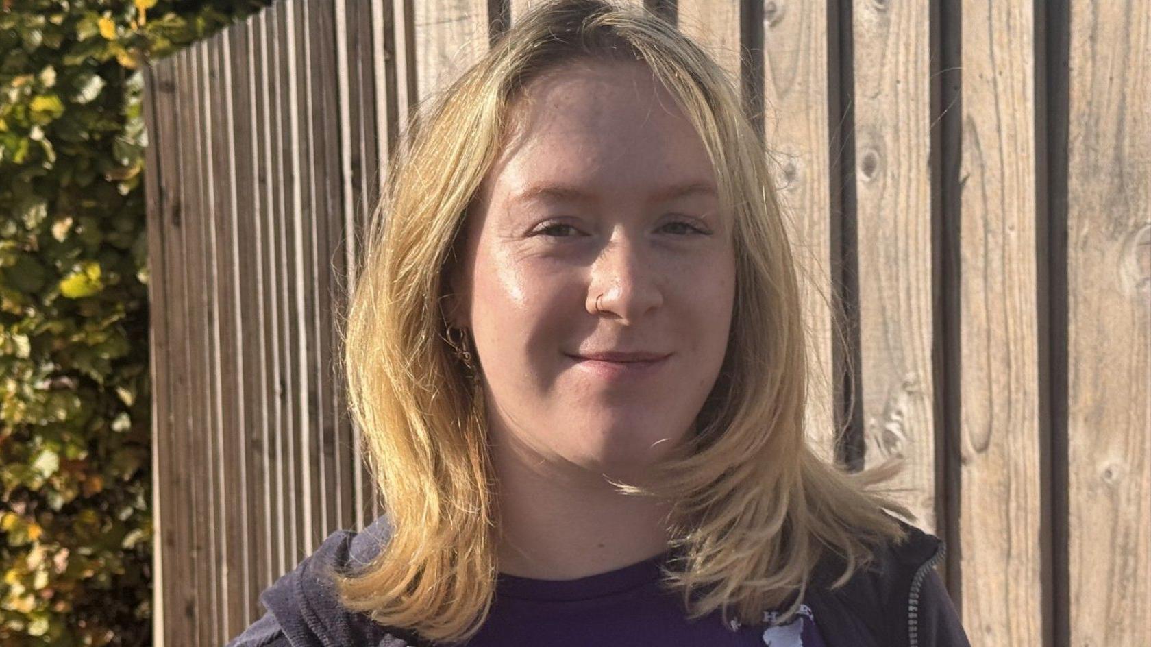 A blonde woman is smiling at the camera. She has shoulder length hair, and is wearing a navy hoodie over a navy t-shirt. She has a nose ring, and is standing in front of a tall wooden fence.