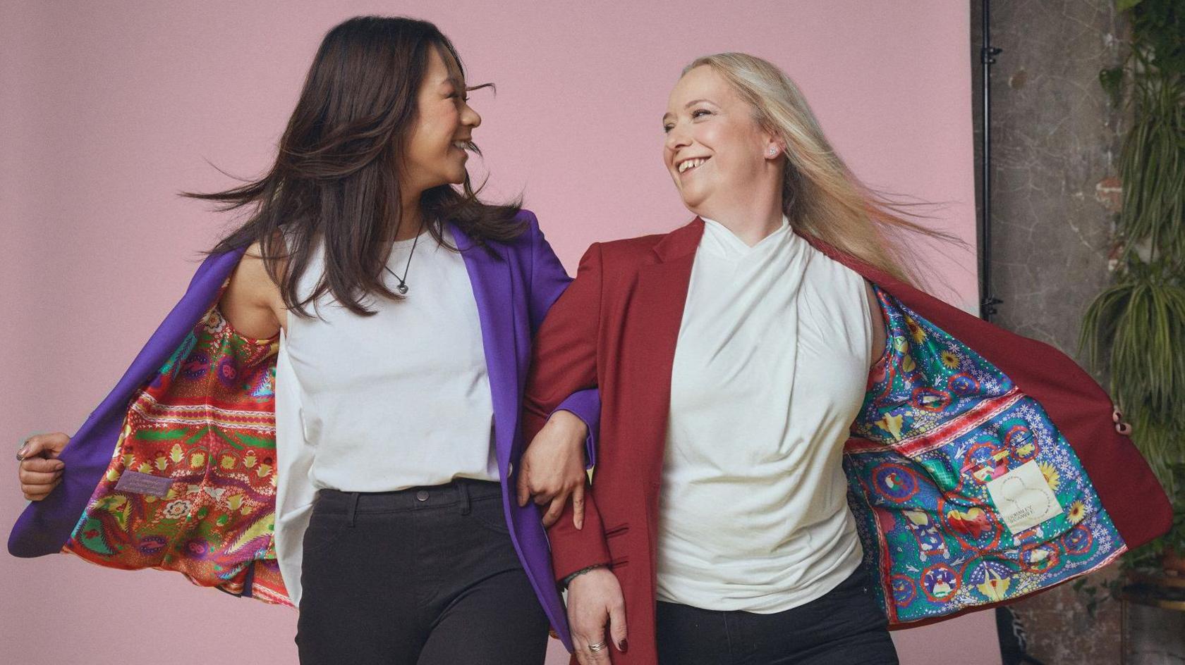 Two women link arms and smile at each other. The woman on the left wears a white top, black jeans and a purple jacket with a colourful lining. The woman on the right wears a white top, black jeans and a burgundy jacket with colourful lining.