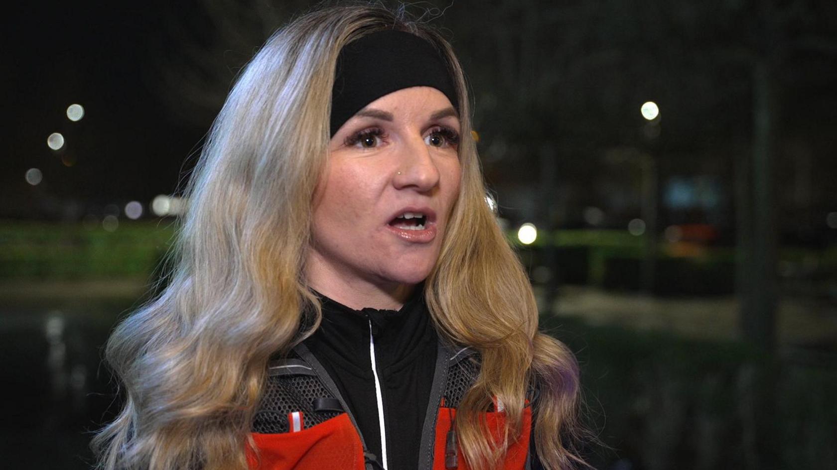 Emma Moffat has long blonde hair and is wearing a black head band. It is night and she is on a street. Emma is wearing a black zip-up top and an orange reflective. Behind you can see a dark street with an area of grass and street lights. 