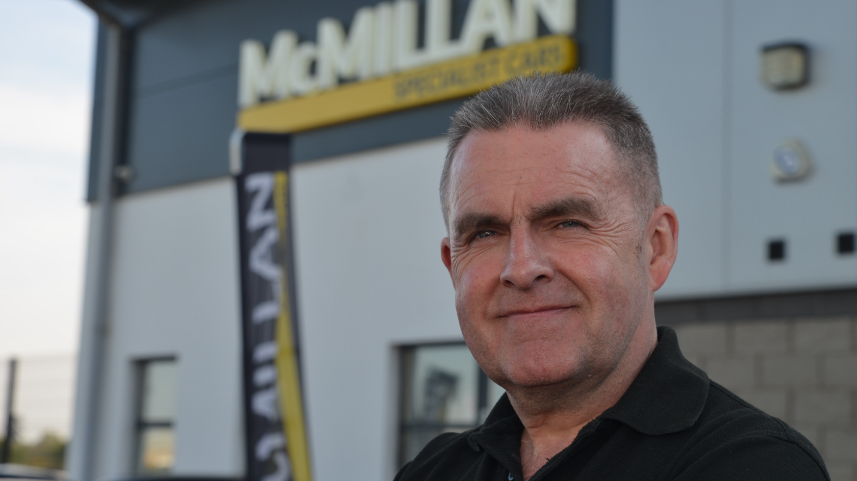 Mr McMillan stands in front of a white and black building with a company logo in the top corner. He has short dark hair and is wearing a black polo shirt as he smiles at the camera.