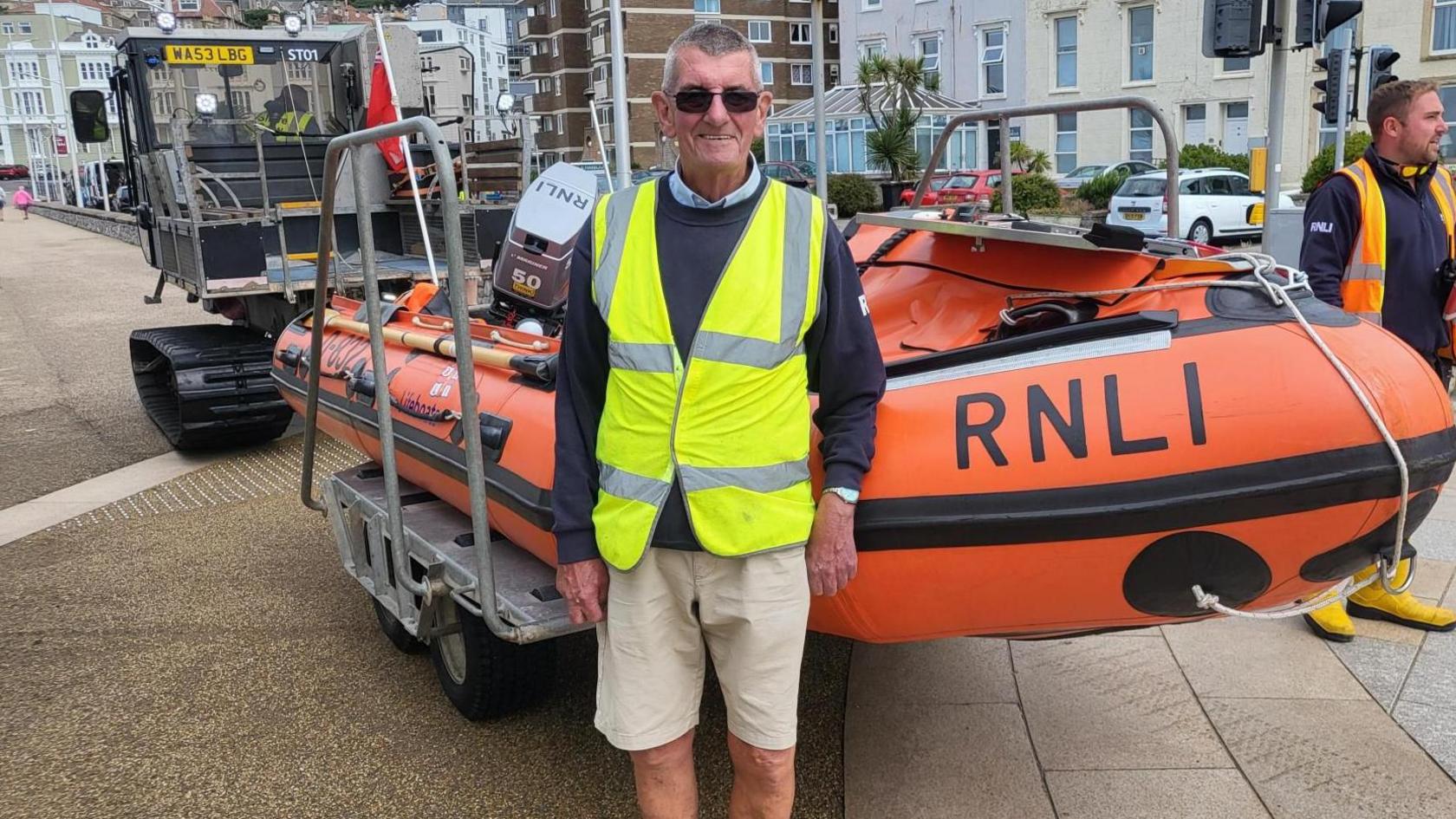 Richard is standing in front of a RNLI speedboat being towed by a truck. He is wearing a dark jumper, beige shorts, a hi-vis jacket and sunglasses.