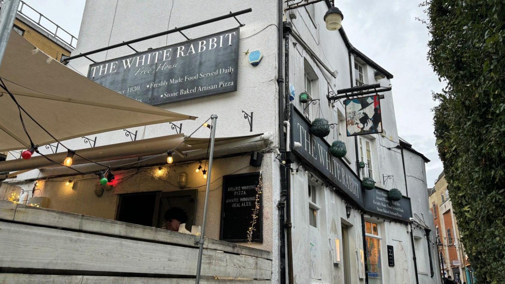 A picture of the outside of a pub called the white rabbit. The pub is white, in central Oxford, with some outdoor seating in the picture. 