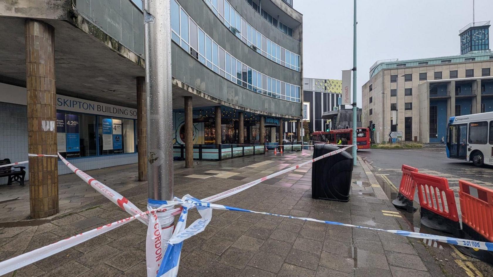 Police tape cordoning off an area of Plymouth city centre. The tape on the right is blue and white and on the left it is red and white. It is tied across lamp-posts and beams. In the distance, on the left, is the entrance of Turtle Bay