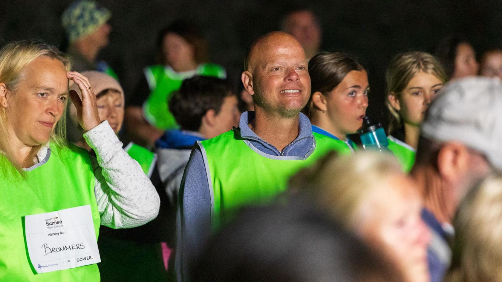 Dressed in lime green tabards walkers wait in the darkness for the start of the walk. One man in the centre of the image is smiling as he looks ahead.