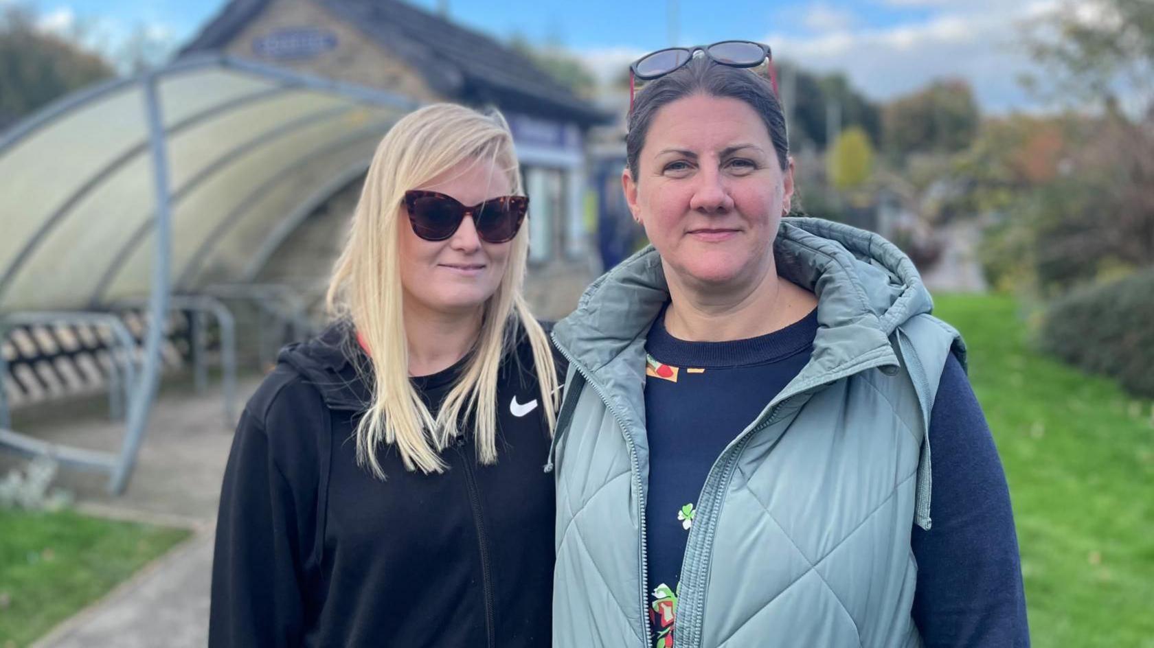 Two middle aged women face the camera. In the background is a bike shed at the train station