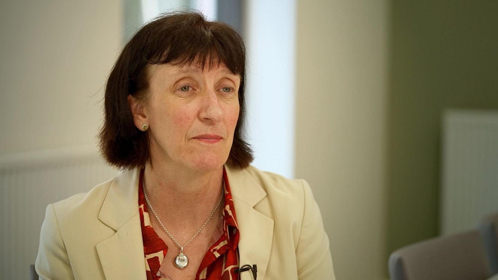 Durham County Council's director of public health Amanda Healy is sitting in a room with walls painted in muted pastels of while and pale green. The arm of a grey chair is out of focus behind her and to her left. She has chin-length dark hair with a fringe and is wearing a cream suit jacket over a red and cream shirt. She has a silver locket around her neck.