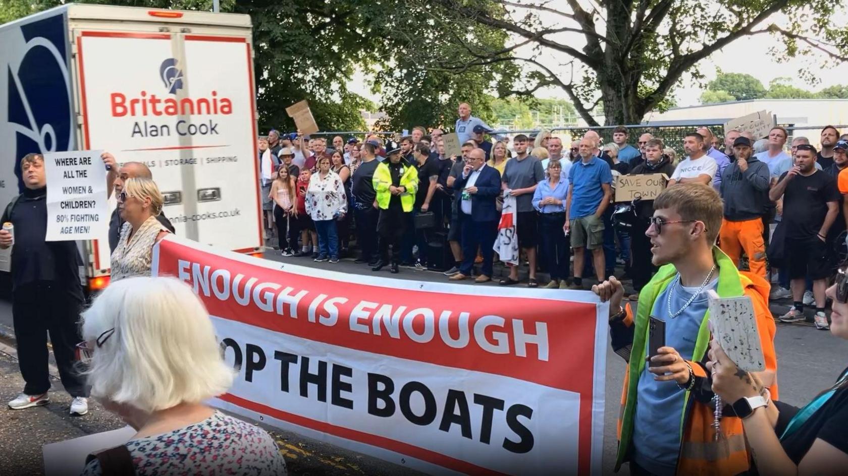 The protestors lined up along a pavement, with more in the foreground. A man and woman hold a large red and white large saying "Enough is Enough, stop the Boats". Another man holding a can in one hand holds up the sign in the caption about women and children. A police officer stands among the crowd. Another placard on a piece of cardboard says "Save our Town".