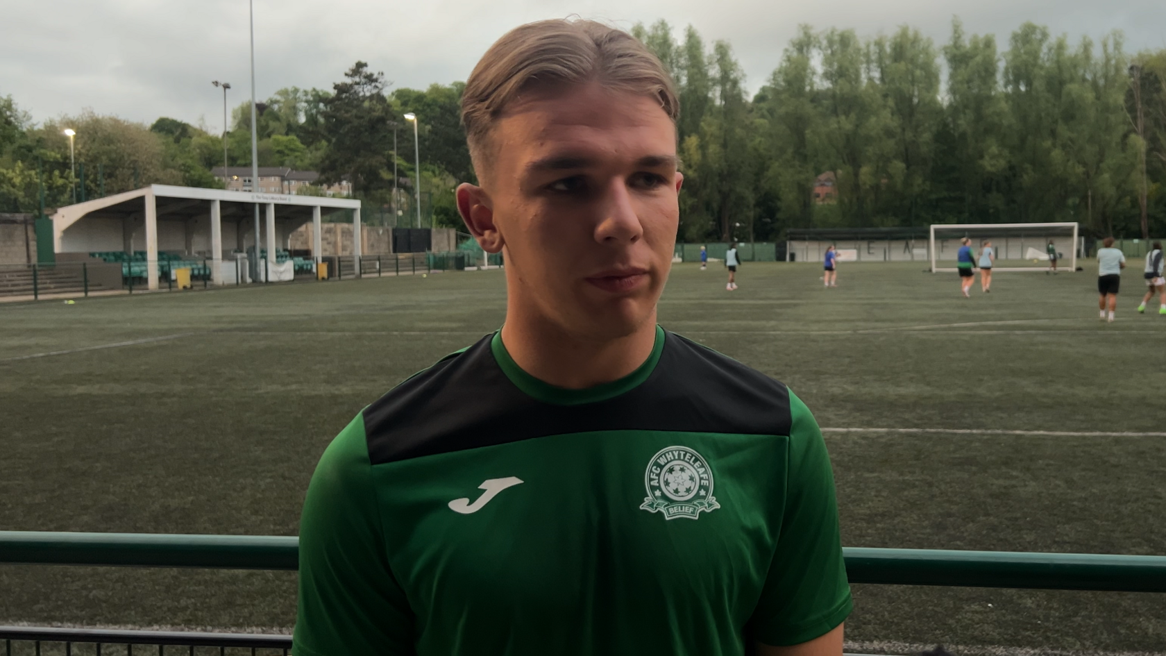 A blonde boy with a centre parting. He is wearing a green AFC Whyteleafe training shirt and is standing in front of a 3G pitch.