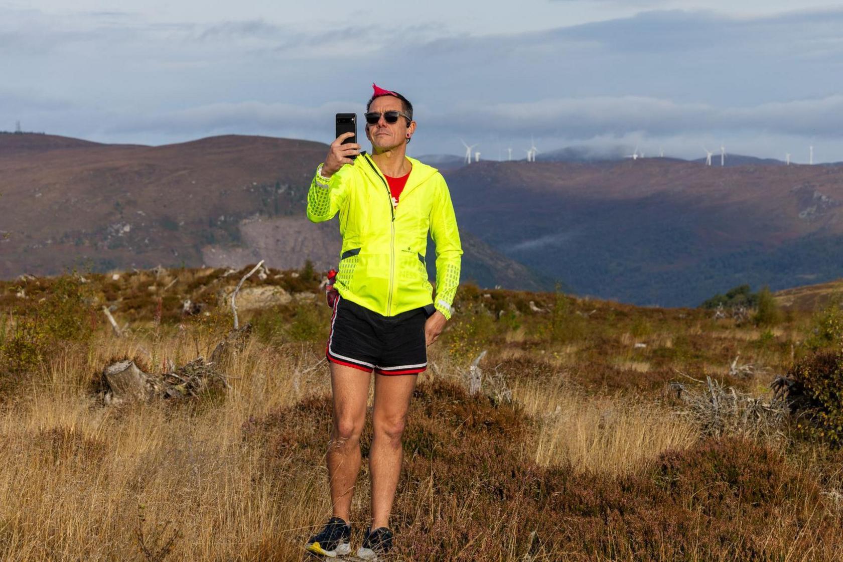 A runner looks at a mobile phone at the start of the Loch Ness marathon. Behind the runner are hills with wind turbines