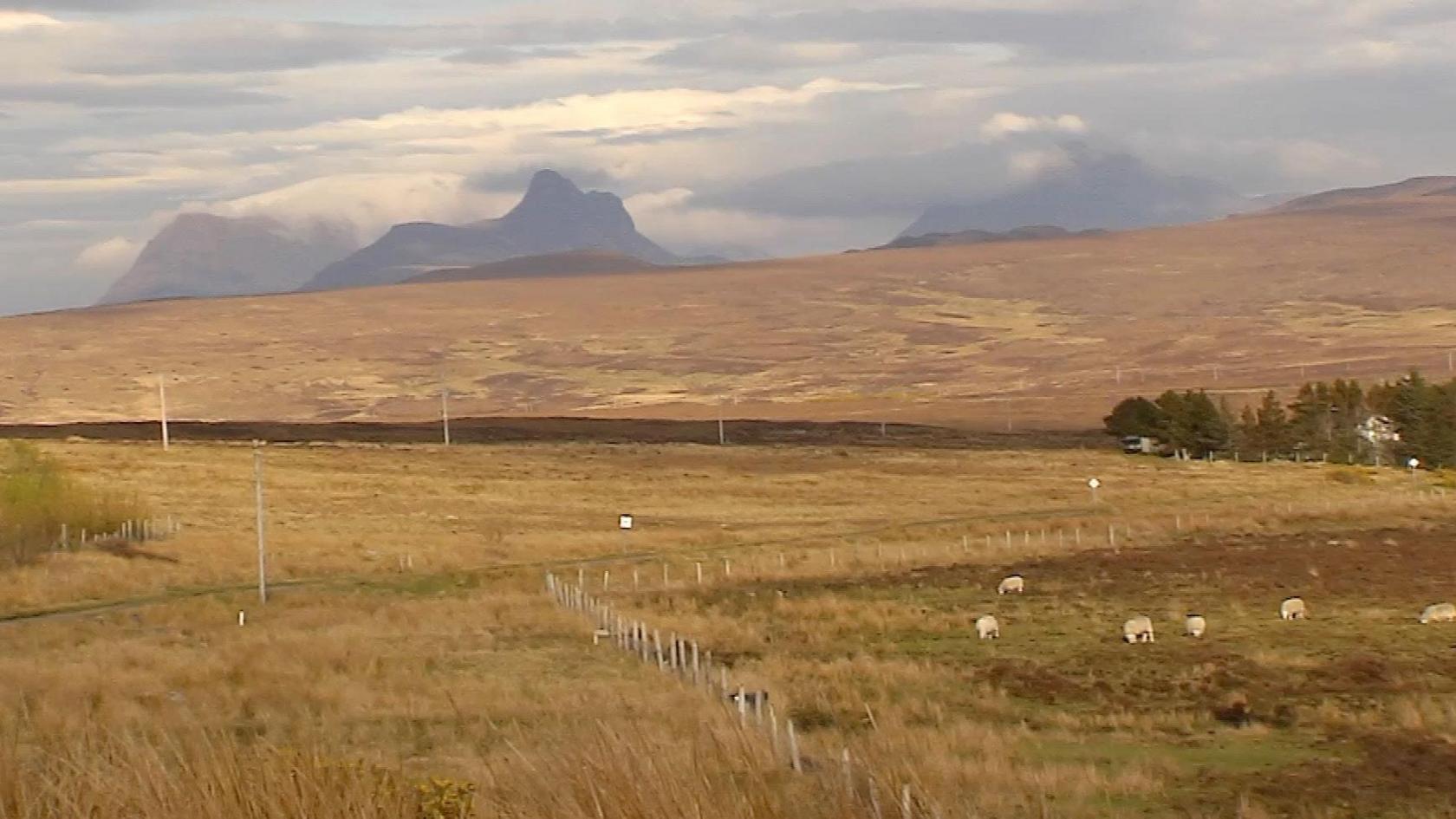 Some of the land now in community ownership. There are hills and mountains covered in cloud in the distance. Sheep are grazing in a field in the foreground.