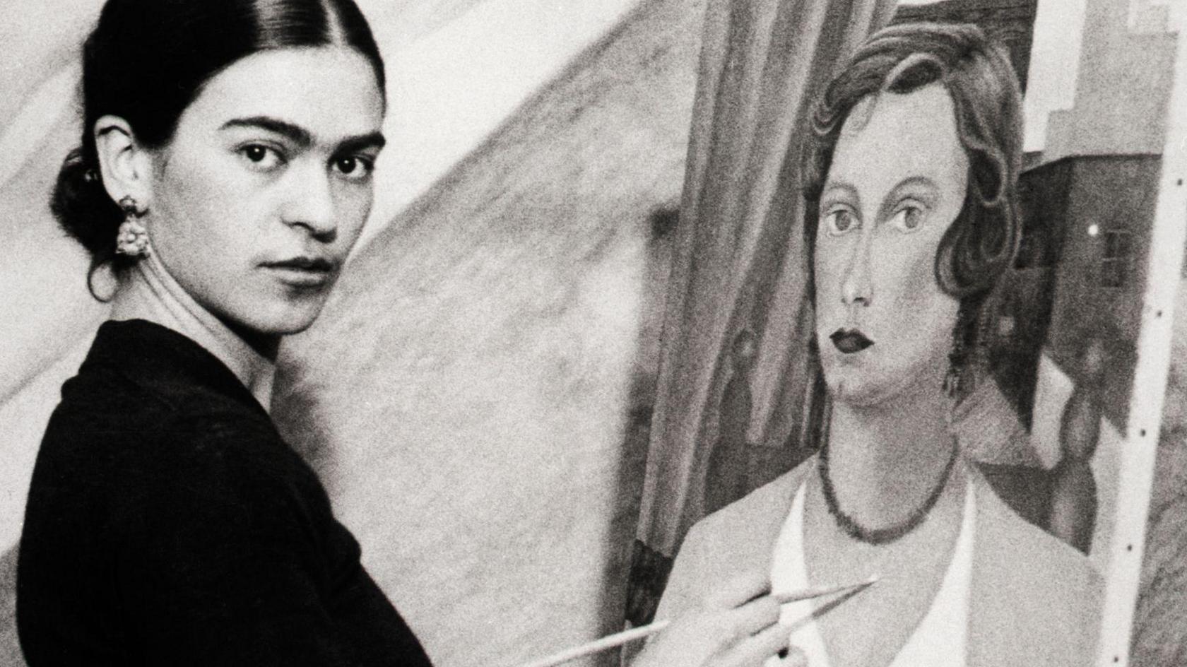 A black and white photo of Frida Kahlo looking directly into the camera, while painting the Portrait of Mrs. Jean Wight.