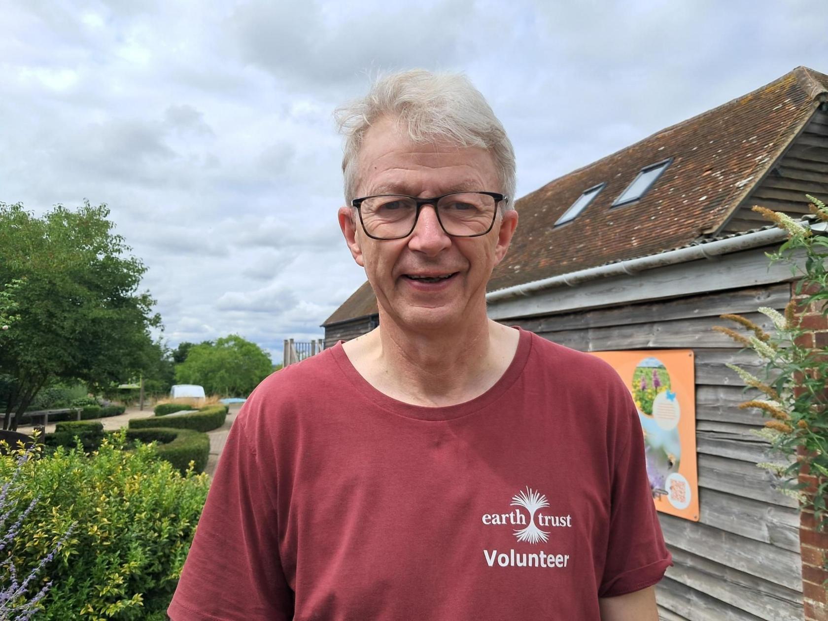 A man with grey hair smiles at the camera. He is wearing black glasses and a red Earth Trust volunteer t-shirt.