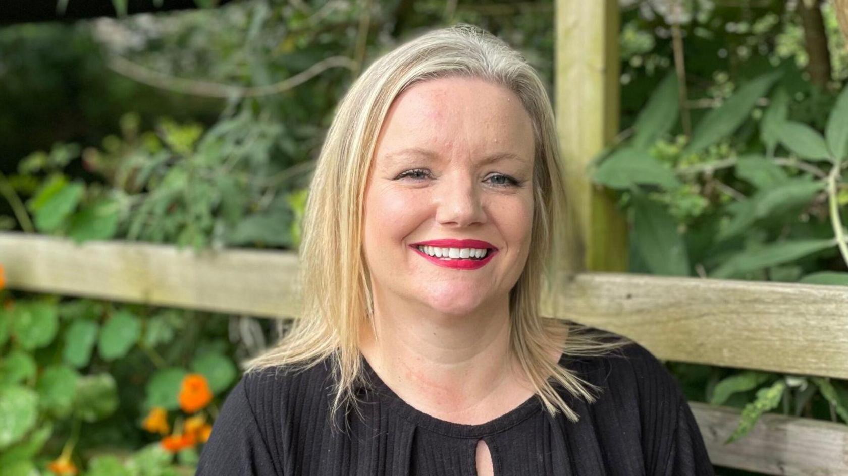 Leigh Yates is sitting on a wooden bench smiling with red and orange nasturtiums in a planter behind her. She is wearing a black top and has long blonde hair,