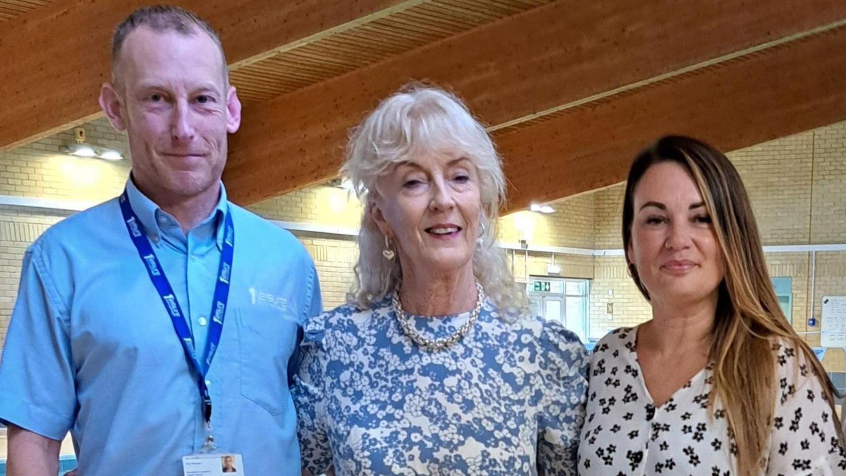 A man wearing a blue shirt and a lanyard, a woman with white hair wearing a blue floral dress and a woman wearing a white floral dress with brown hair stand with their arms around each other