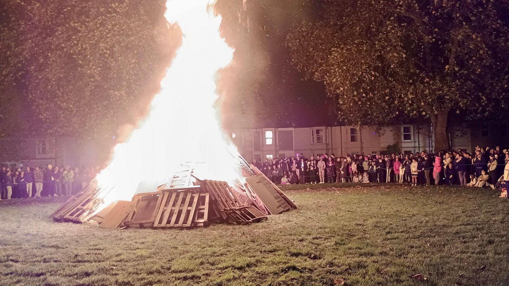 A group of people gather on the edges of a green near houses to watch a bonfire made using wood and old pallets