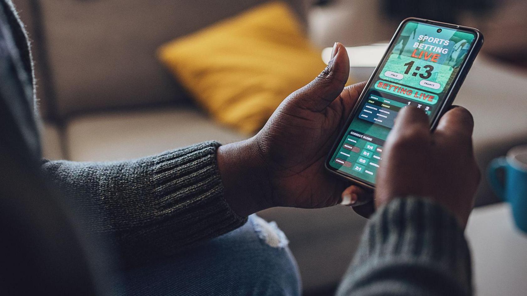 A man looking through a 'sports betting' website on his smartphone. He has blue jeans on and a green jumper and is sitting on the sofa. His face is not in the image.