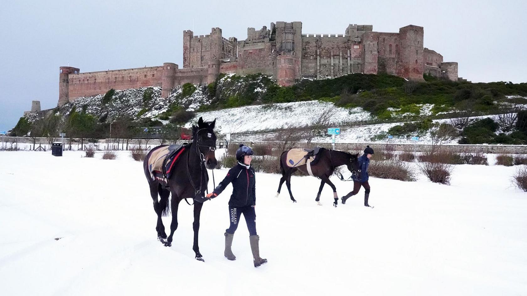 A woman walks with a horse in front of Bamburgh Castle in Bamburgh - the ground is covered in snow