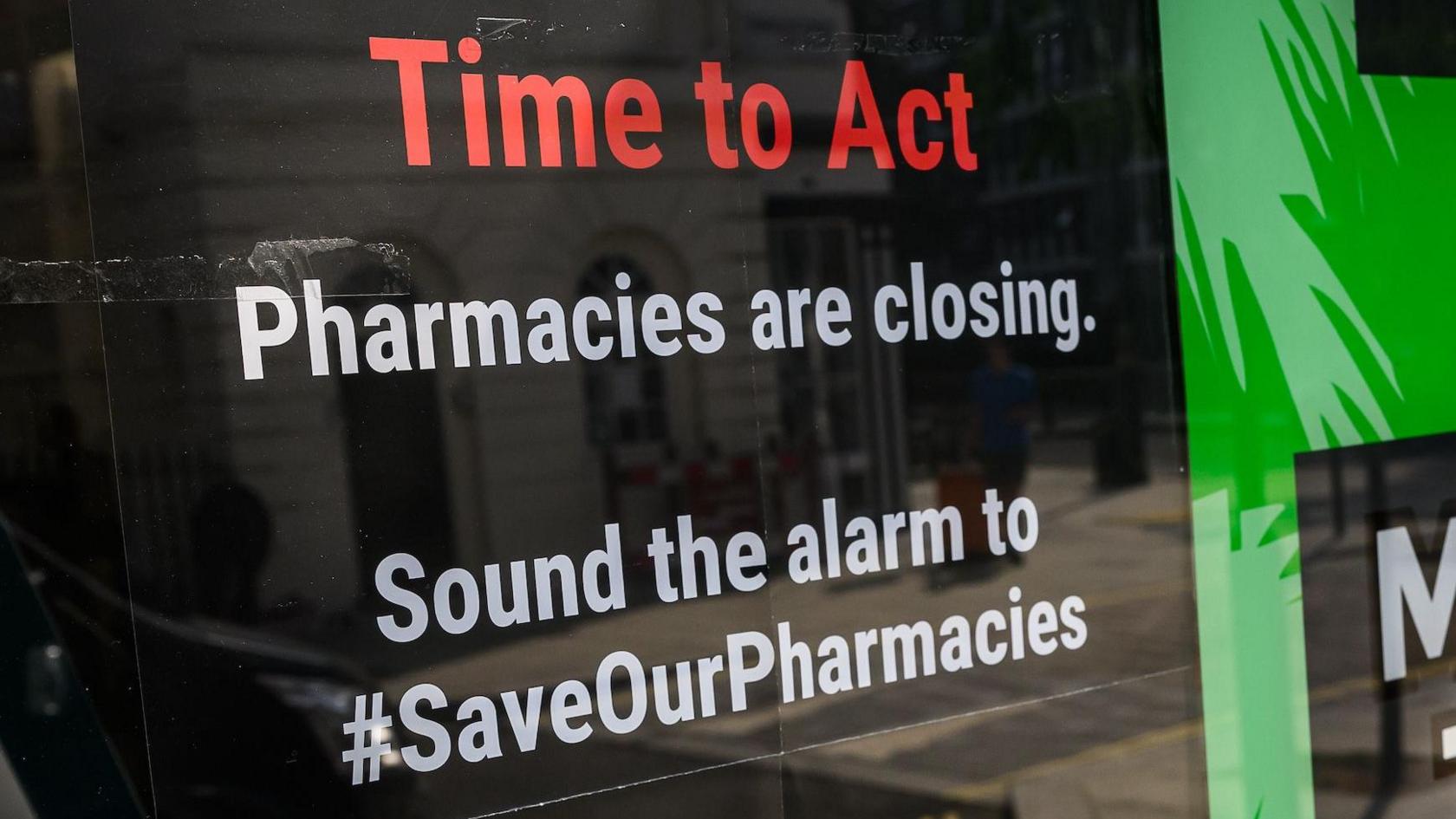 Two posters in the window of a pharmacist branch. The first is on a black bakground with white lettering that reads, 'Pharmacies are closing. Sound the alarm to to save our pharmacies. The second says 'Malaria tablets; against a green background with a graphic of palm trees.