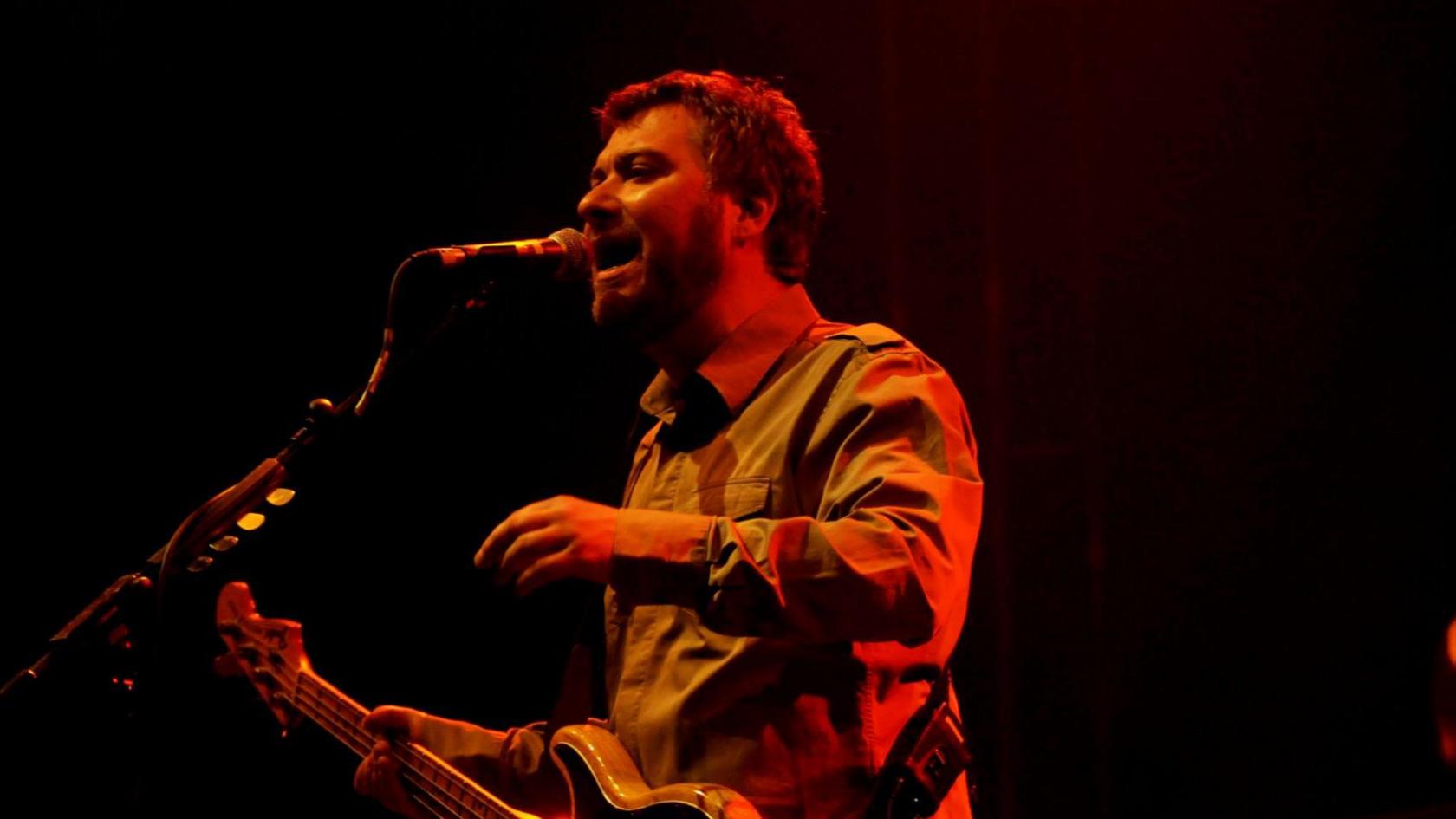 Jimi Goodwin playing bass and singing into a microphone on stage wearing a khaki shirt against a blacked out backdrop. 