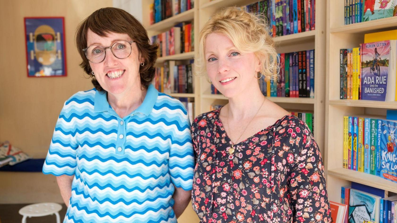 Two women standing in a bookshop. One woman has short brown hair, glasses, and a blue and white t-shirt. The other woman has blonde hair and is wearing a black and red floral dress.