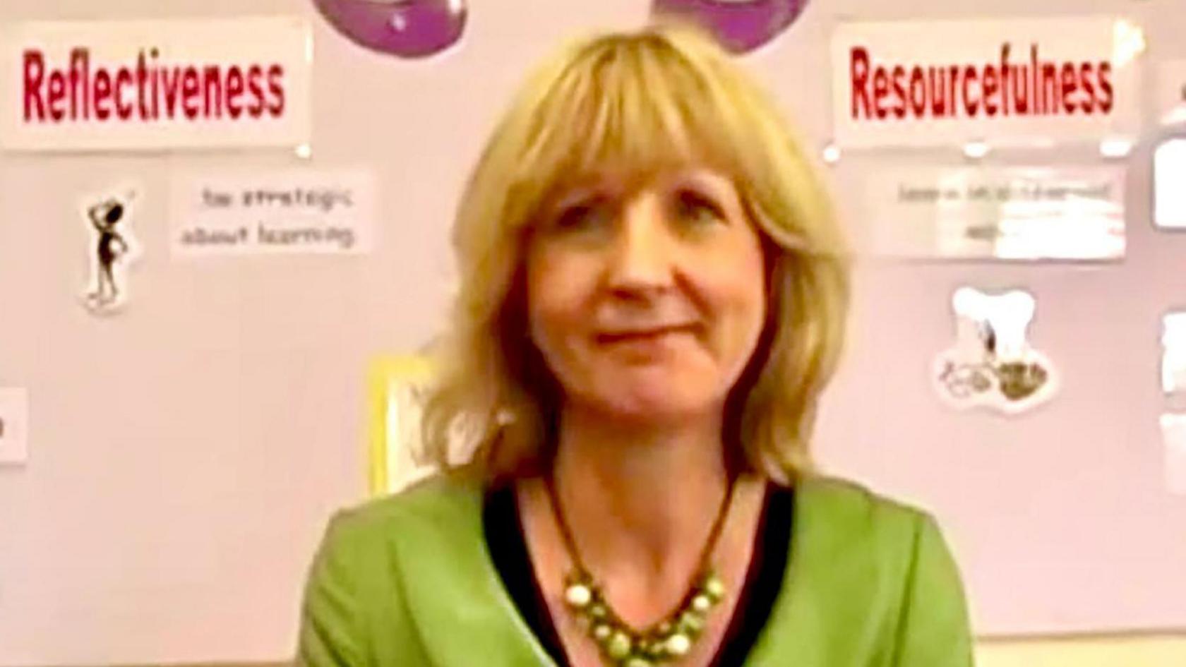 Christine Baker is seen in front of a school noticeboard showing a display about learning strategies. She has blonde hair with a fringe and is wearing a green jacket with a black top and a necklace of green beads.
