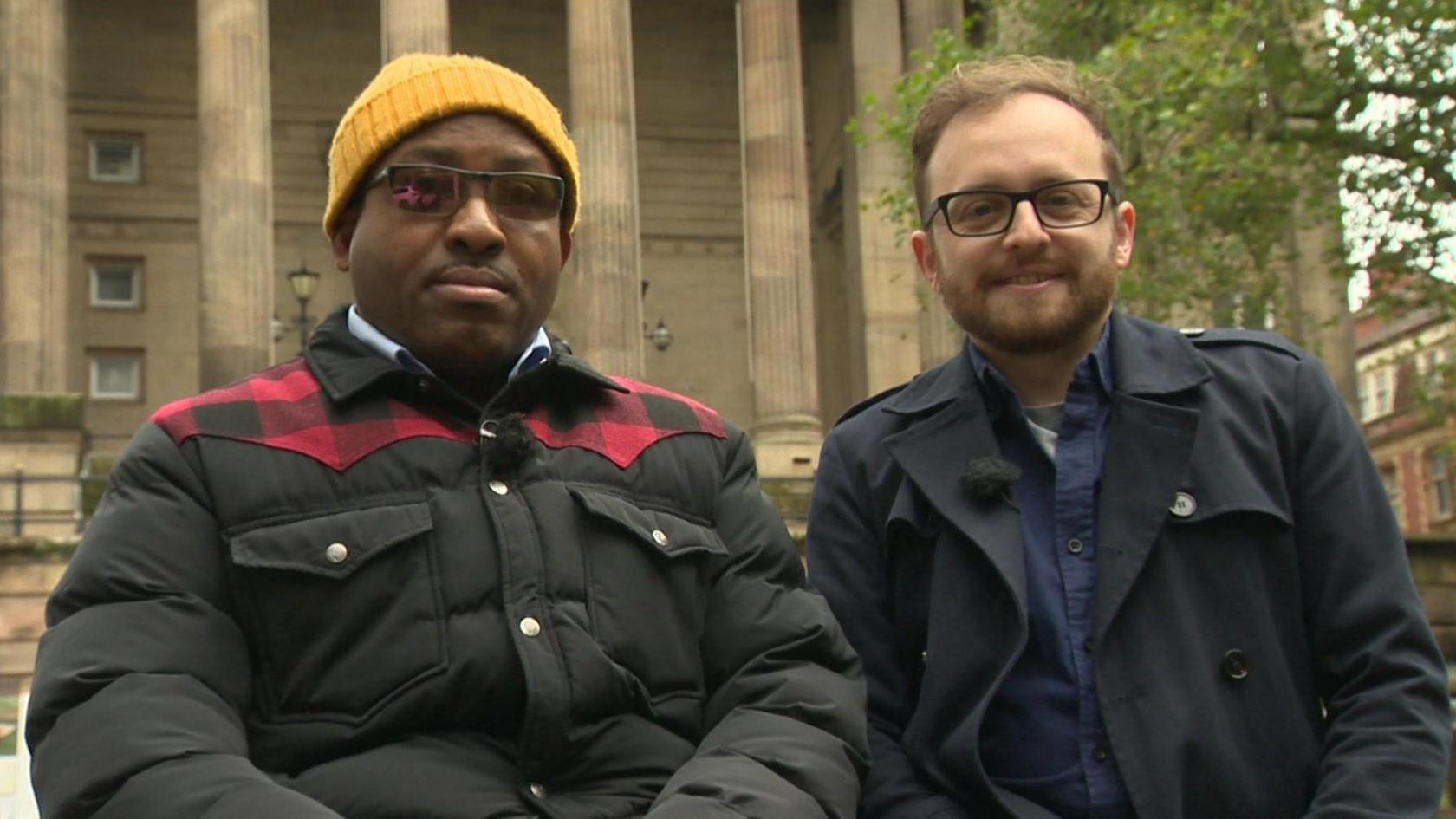 Two men, wearing winter coats, sitting on bench in a square in Preston city centre.