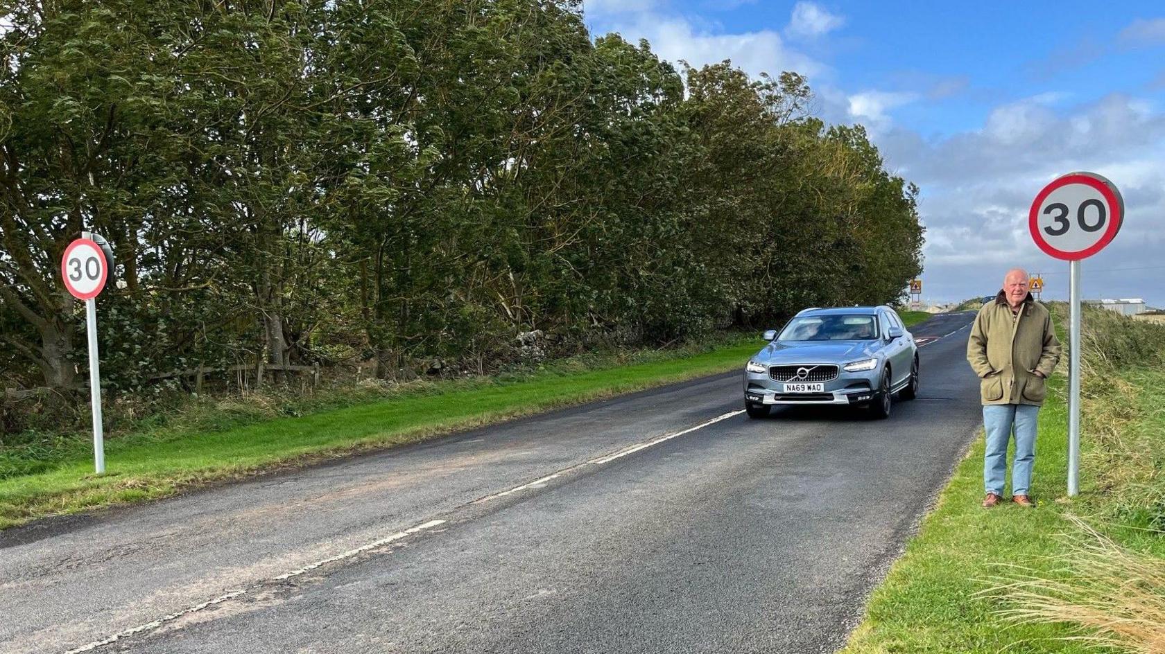 John Rhind, wearing an olive green jacket and jeans, stands at a 30mph sign as a car approaches on a rural road. 