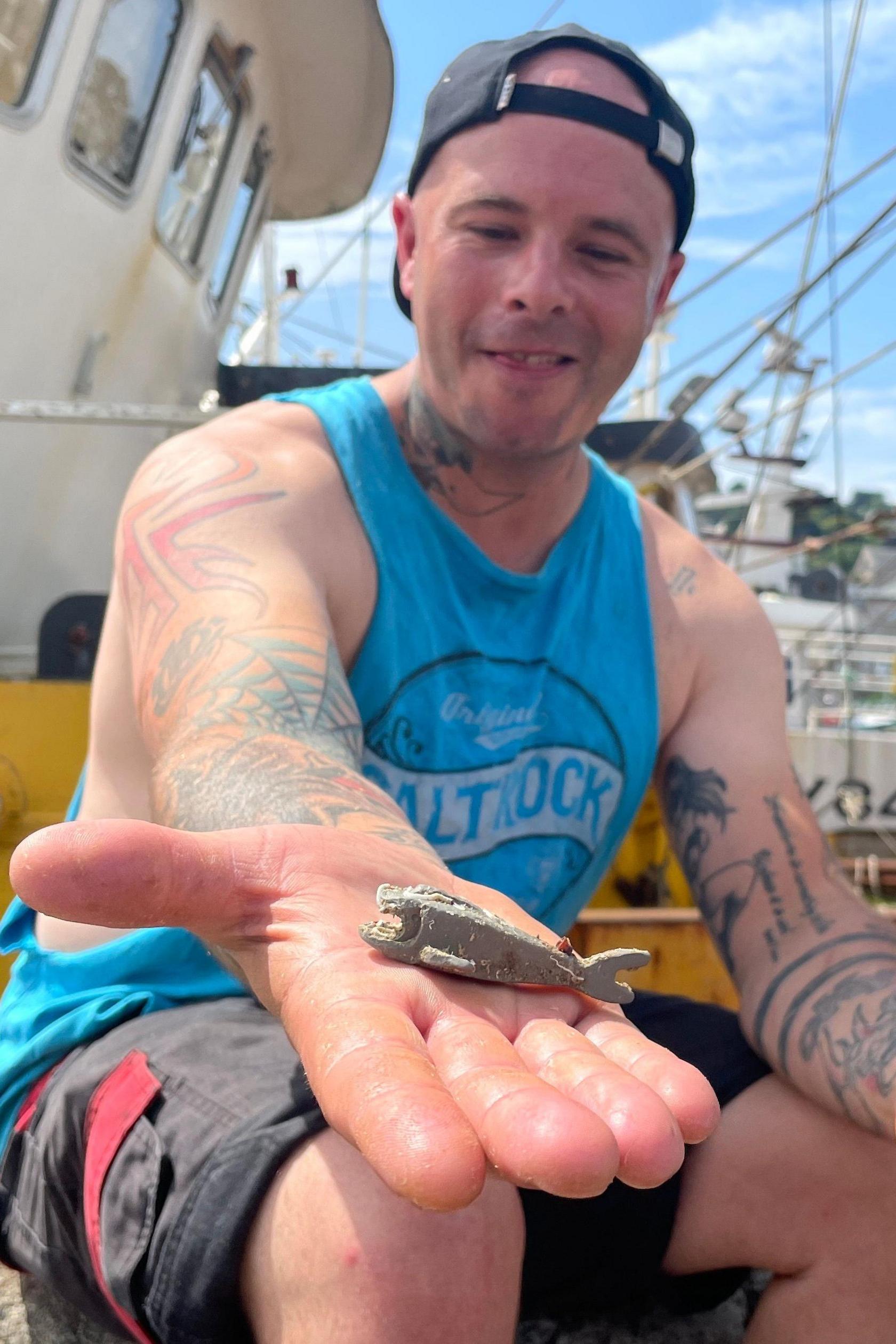 Richard West on the fishing trawler smiling broadly and looking at the grey Lego shark on his open hand. He is wearing a vest top and shorts together with a cap worn backwards.