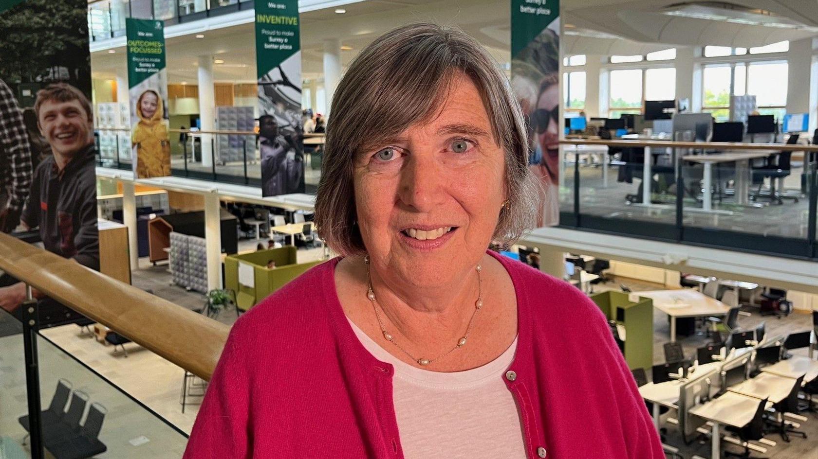 Clare Curran in the foyer of Surrey County Council headquarters. She is wearing a pink top and is standing on a balcony overlooking a room with rows of desks