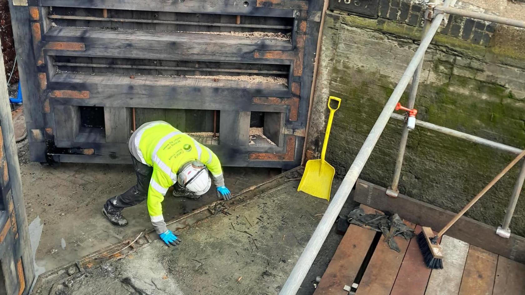 A worker in an orange hi-vis and white hard hat with blue gloves is working at the base of a new lock gate.  He has both hands on the ground. There is a yellow spade propped up against the lock wall.  A wooden walkway is visible to the right.