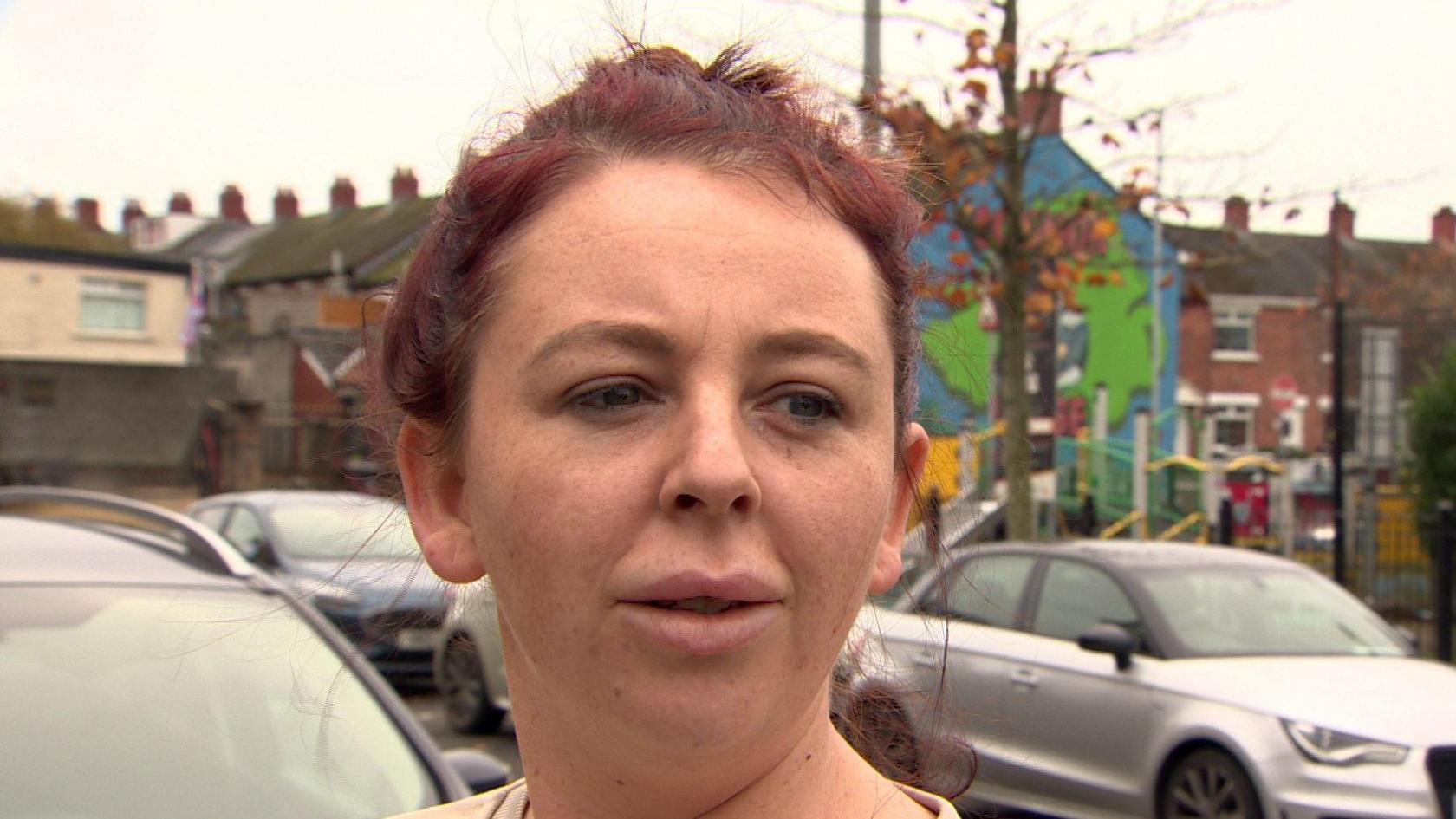 Danielle Pollins, who has red dyed hair tied up and is wearing a cream jumper standing outside a park. 