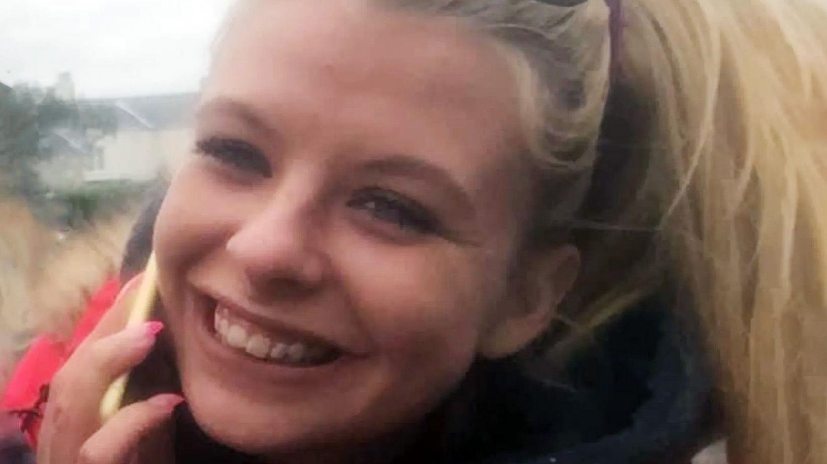 A young woman with blonde hair and a mobile phone held to her right ear smiles at the camera