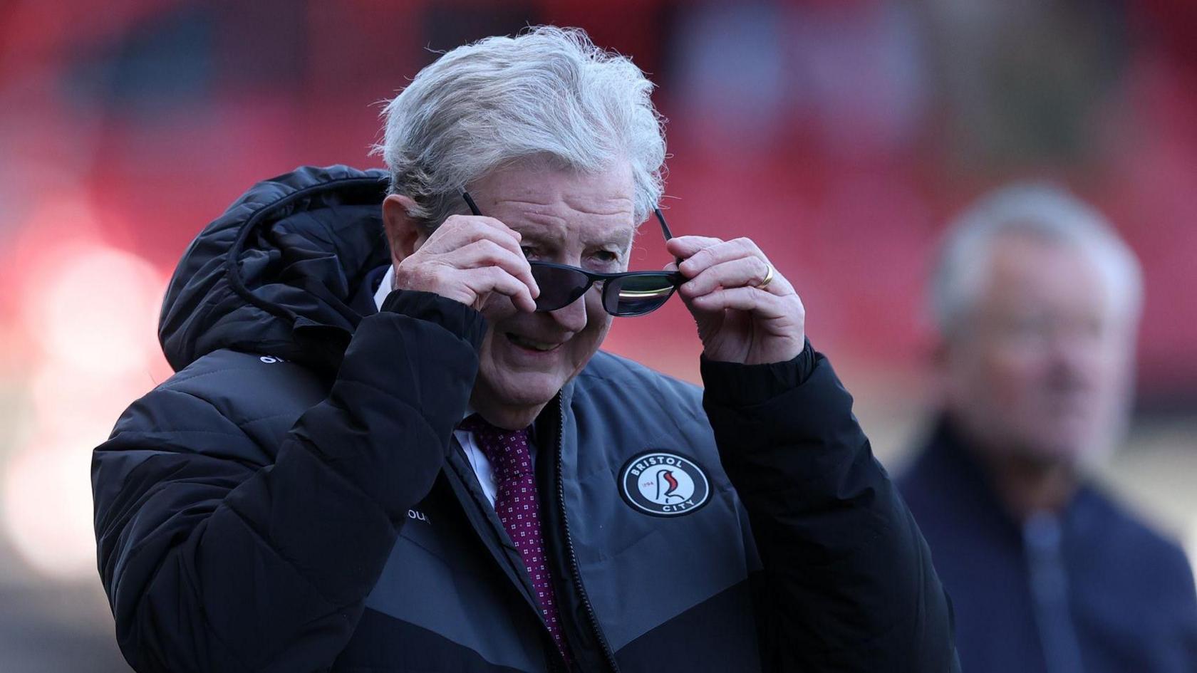 A photo of Roy Hodgson putting sunglasses on while wearing a black Bristol City jacket and a red tie