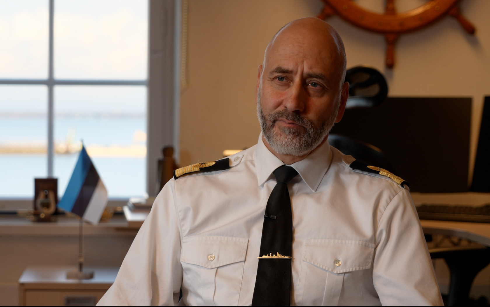 Commodore Ivo Värk wears a white shirt with epaulets and a tie with a pin. Behind him is the Estonia flag on a desk and a ships wheel mounted on the wall behind a desk.