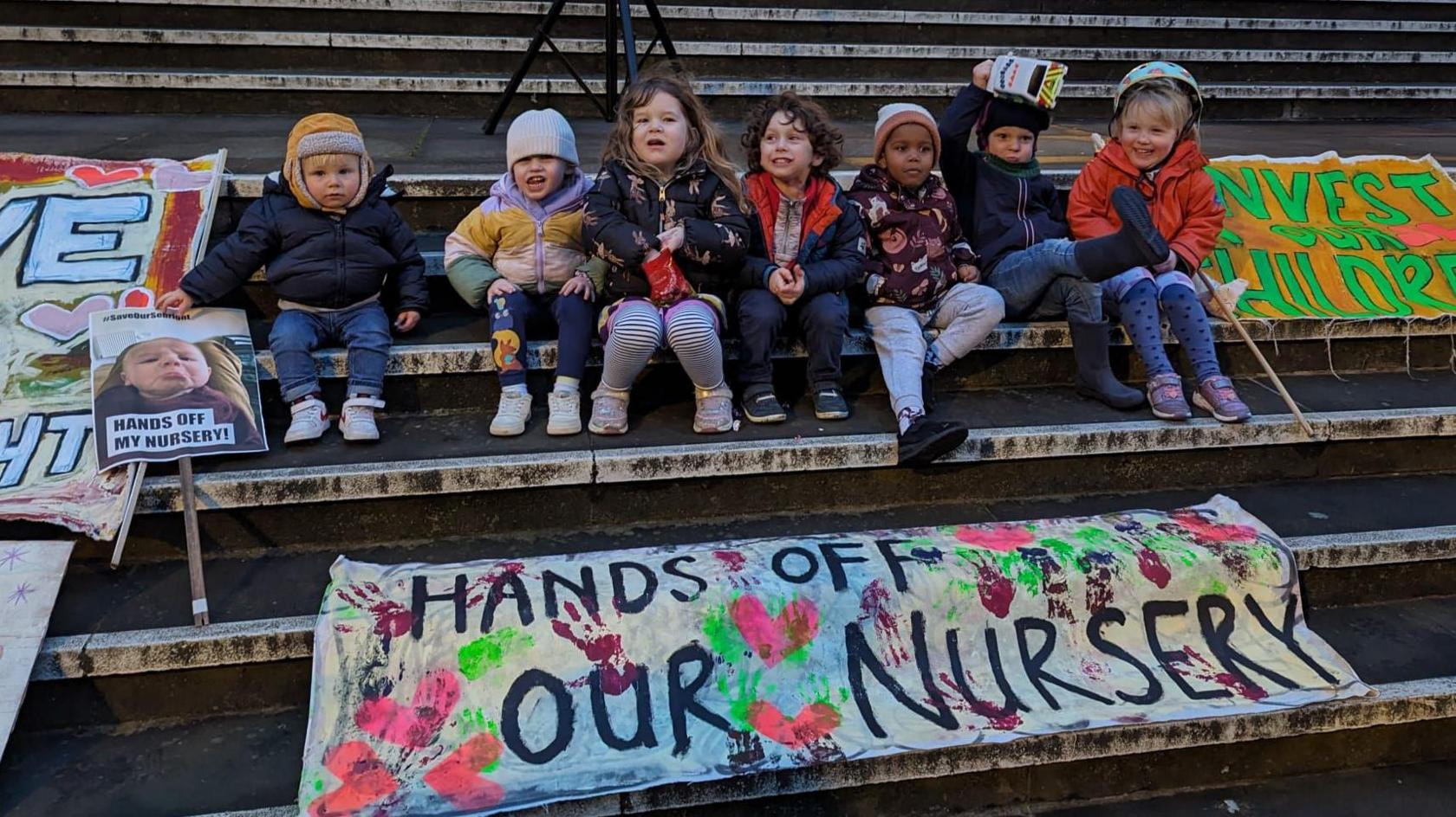 Seven young children sit on the steps of the council offices holding placards saying "Hands off our nursery"