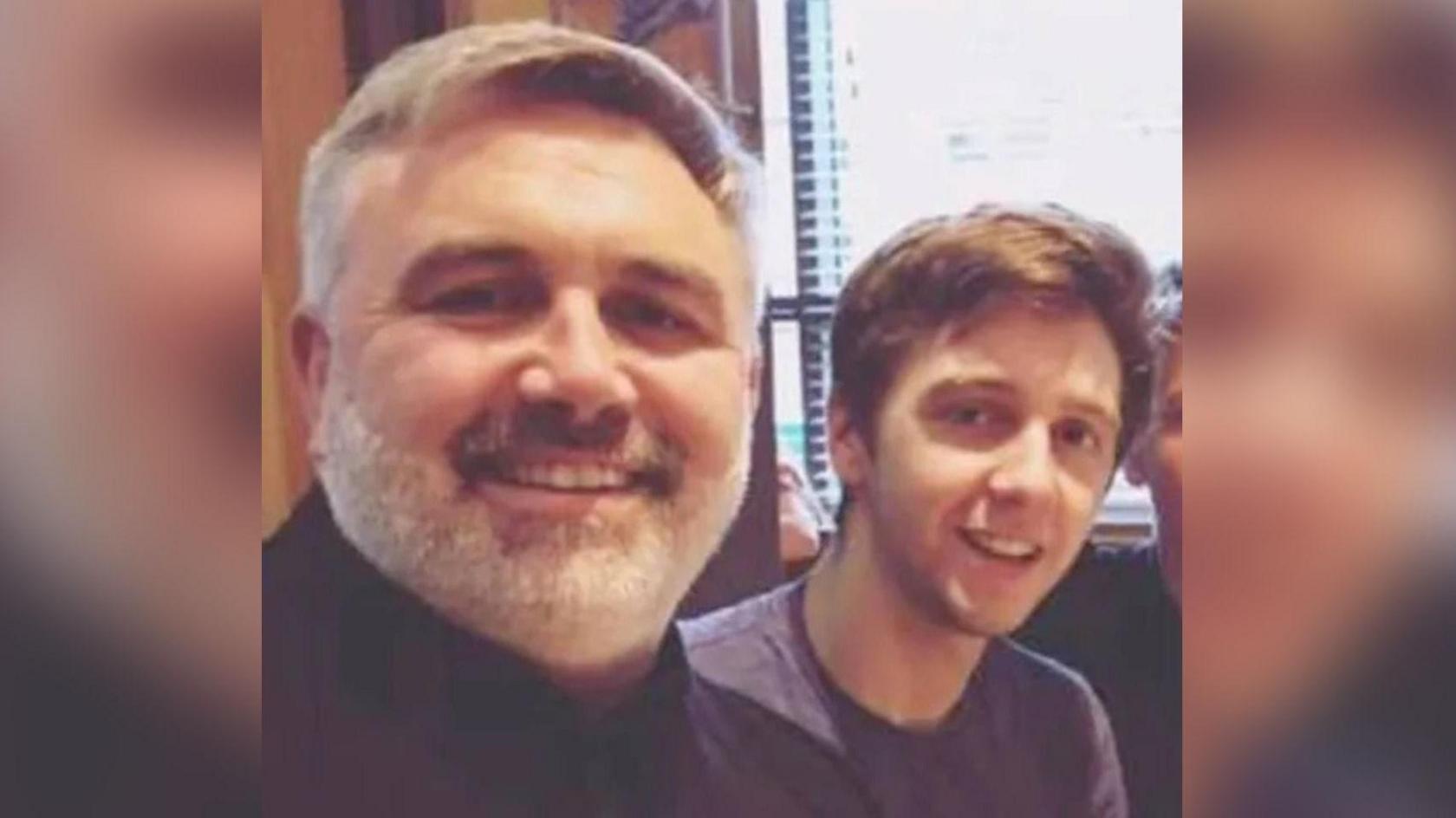 Daniel and Liam both look at the camera and are smiling. Daniel is closer to the camera and has a short grey and white beard. In the background you can just see a window with blinds pulled down.