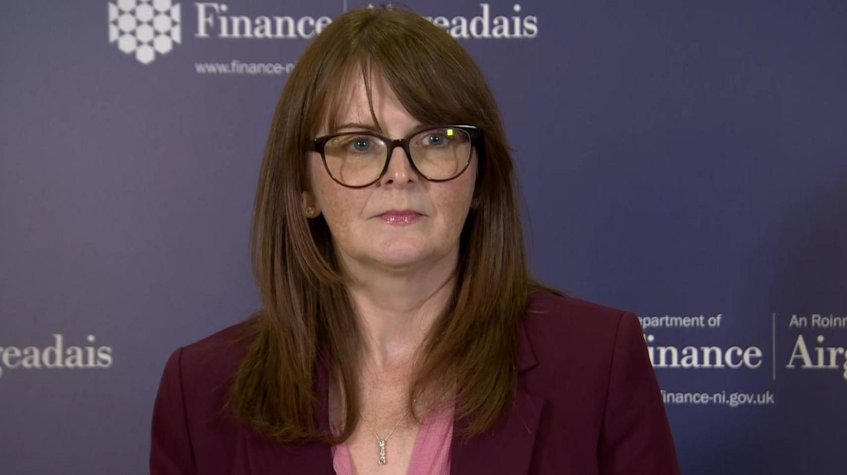 Northern Ireland's Finance Minister Caoimhe Archibald addressing the cameras at a press conference.  She has shoulder-length auburn hair, dark-rimmed glasses and is wearing a pink blouse and a claret-coloured blazer.