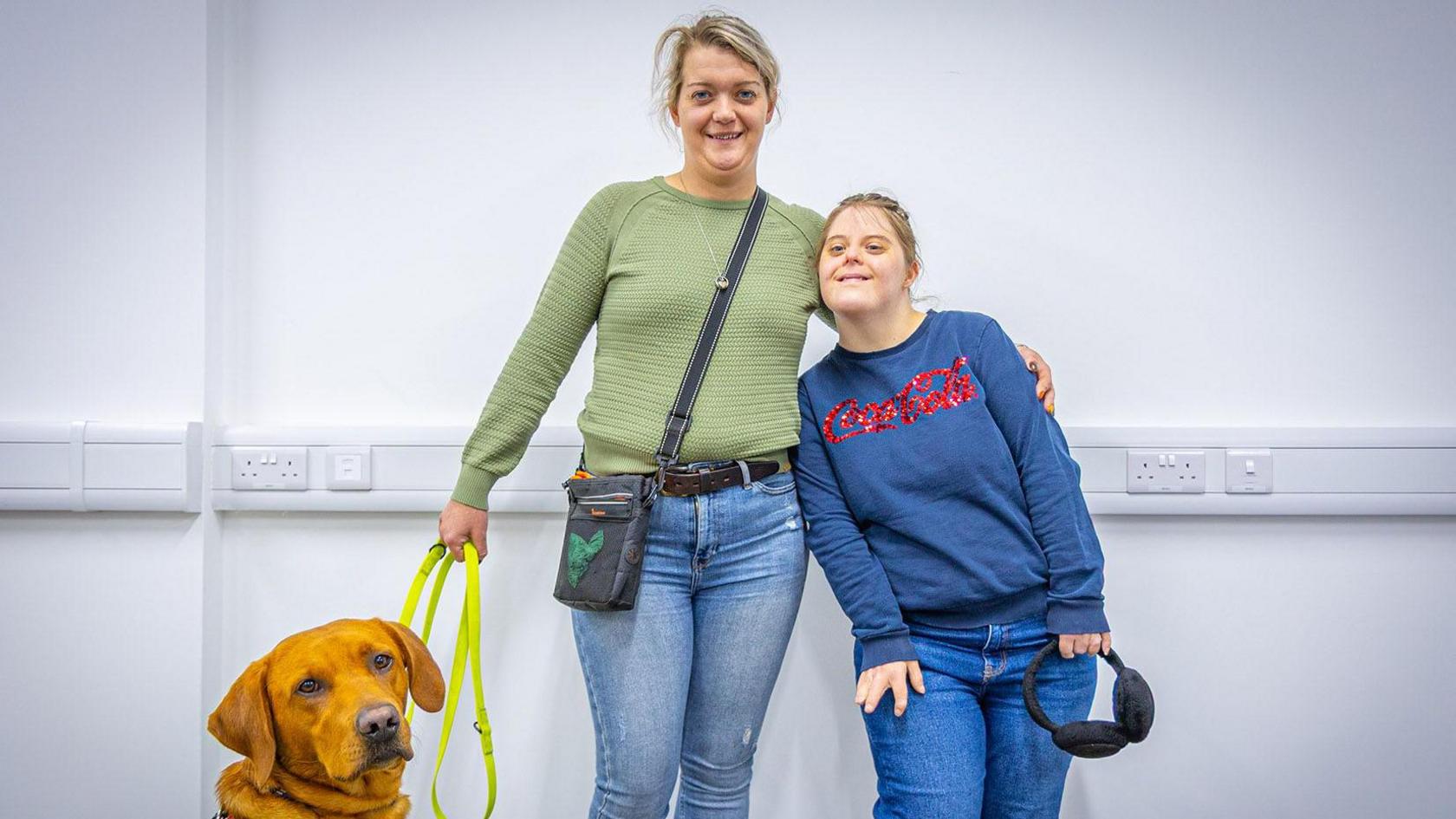 Aynsley is wearing a green jumper with jeans, and has blonde hair tied back. She has her arm around her sister Emma, who is wearing a navy jumper with a coca cola logo on it, and jeans. Aynsley has a ginger Labrador on a lead. 