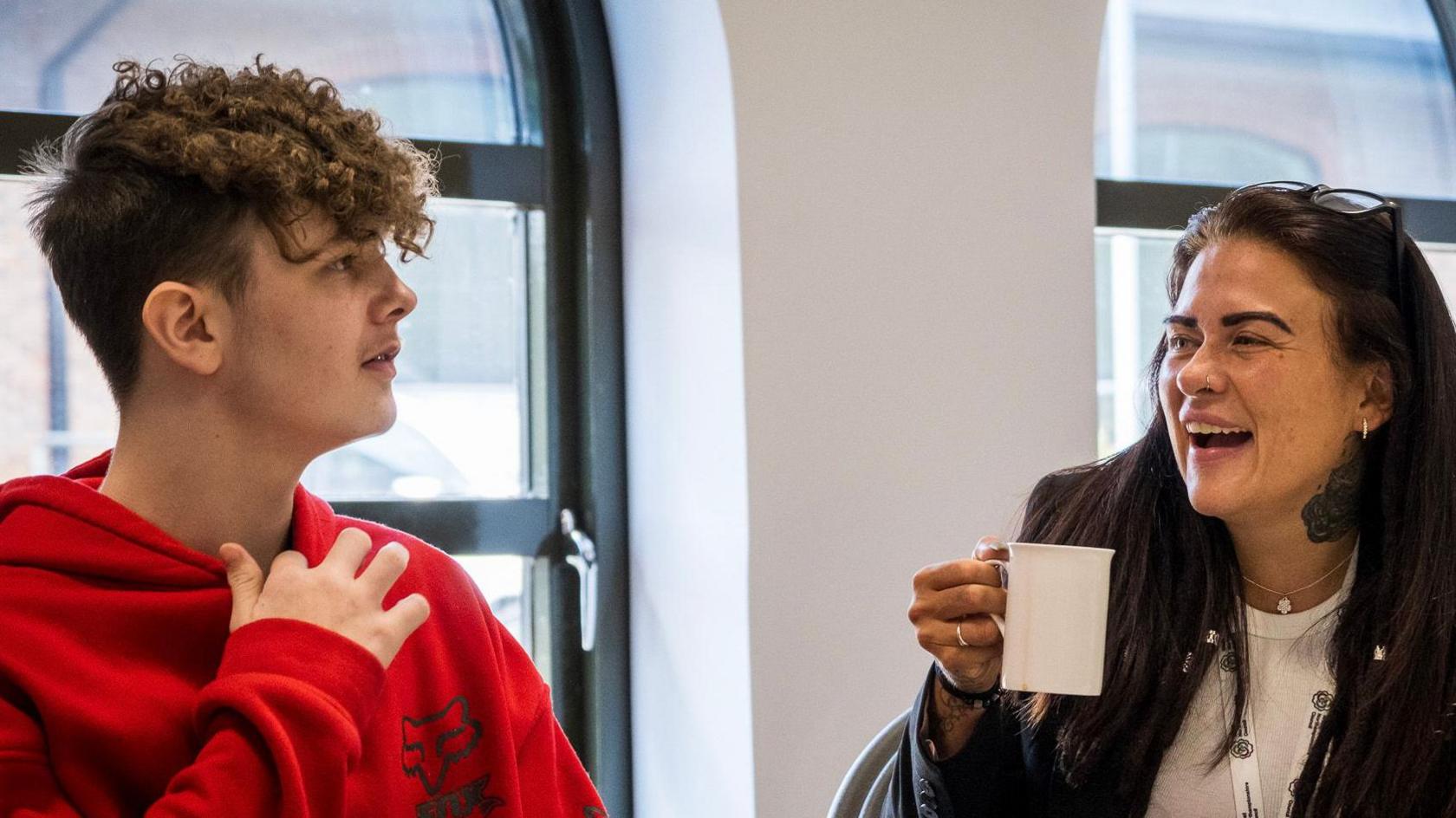 A teenage pupil on the left in a red hoody looking towards a woman in a classroom. He has short mid brown hair, curling on his forehead. The woman, who is to his left, is looking towards him and laughing. She has long black hair, her glasses are resting on her head, she is wearing a black jacket over a white T-shirt and she has a white mug raised in her right hand.