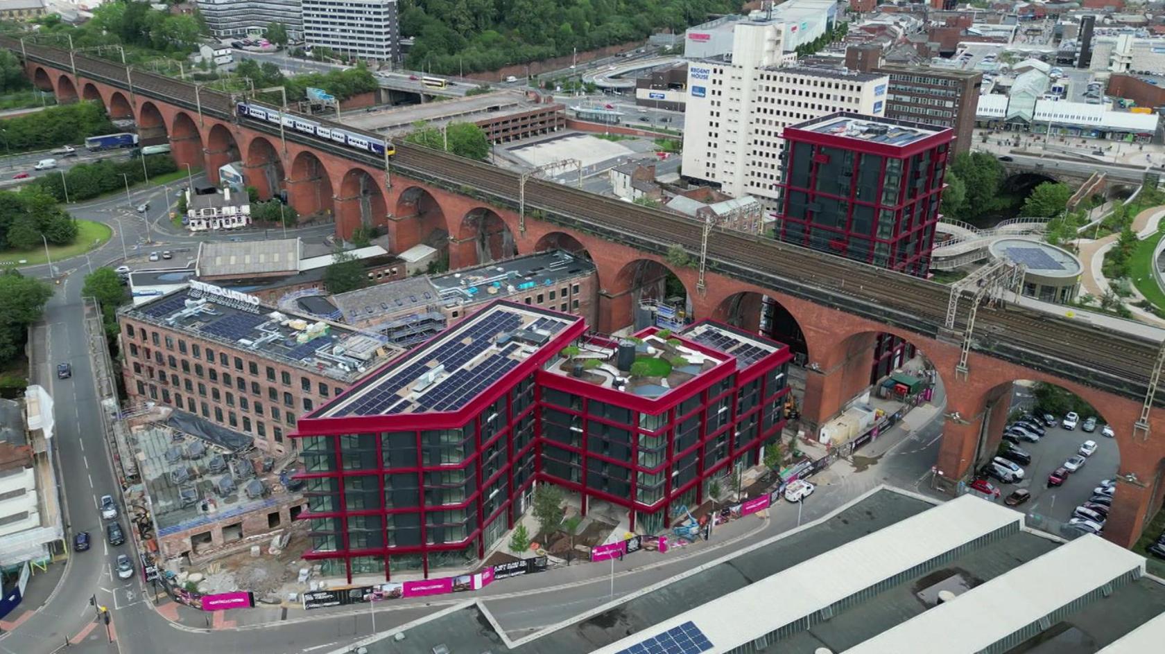 A drone image of Stockport town centre. There is a train travelling along the historic viaduct which spans the length of the picture. In the middle of a ring road is a number of new, modern high rises located next to former mill buildings.