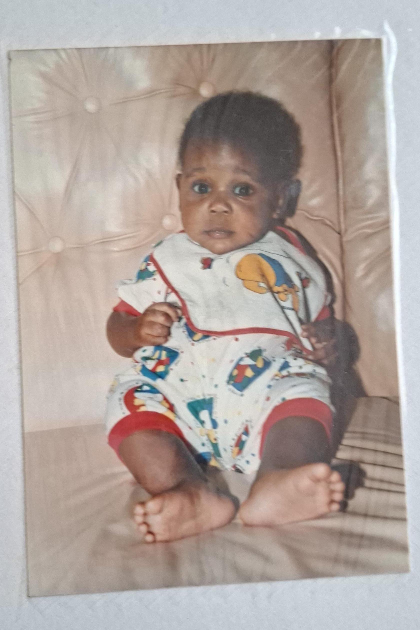 A baby in bright baby clothes and a bib sits on the sofa and looks at the camera.
