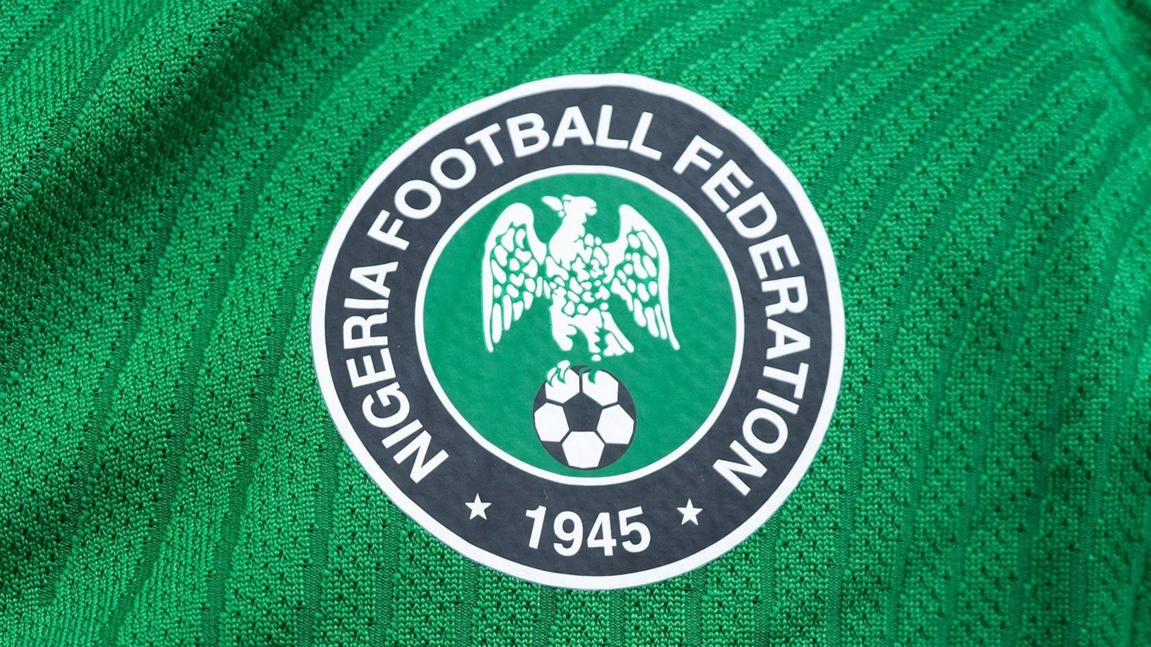 A close up of the Nigeria Football Federation's circular crest sitting on a green shirt. The crest depicts a white eagle perched on a black and white football on a green circular background which is surrounded by the wording Nigeria Football Federation 1945 written in white text on a black background which goes around the outside of the design