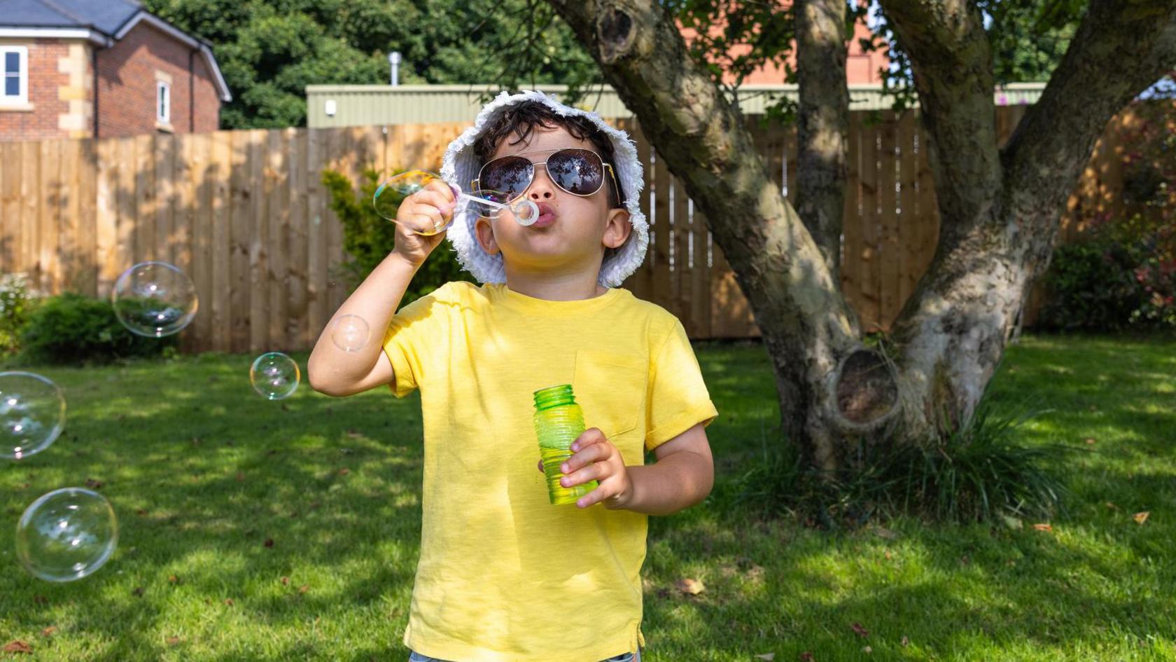 Boy dressed in yellow t-shirt blows bubbles in a garden.