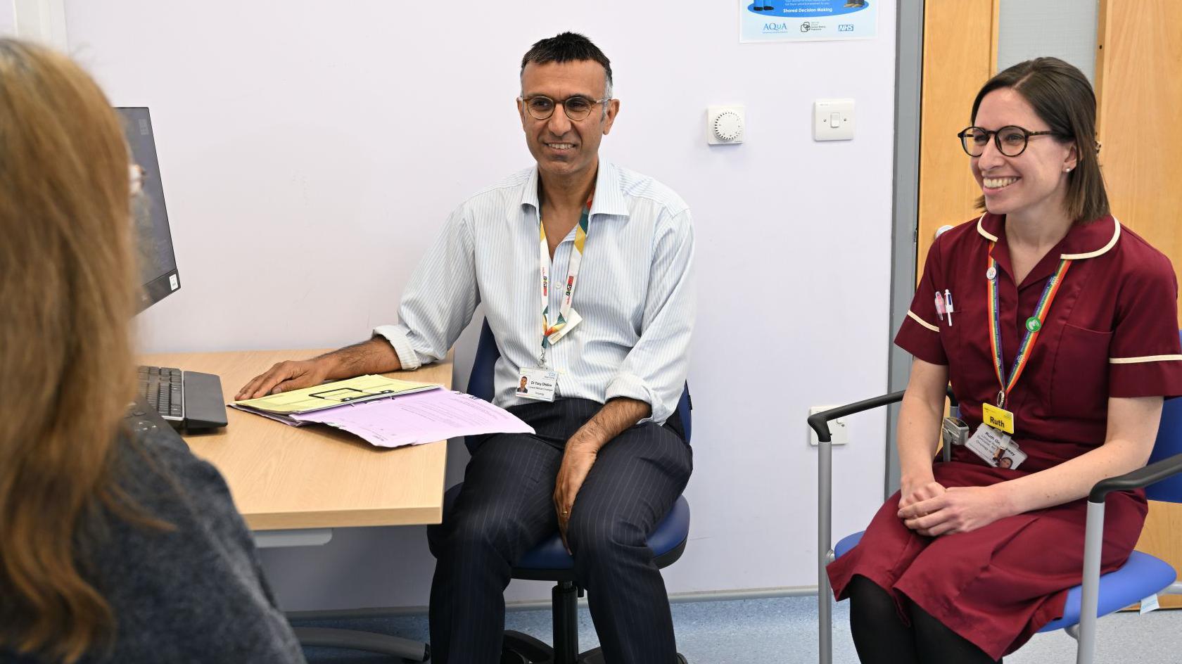 a doctor and nurse speak to a female patient sitting with her back to the camera in a clinical space