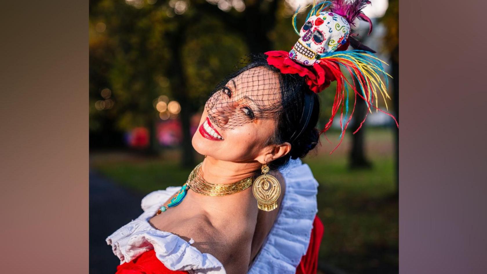 Red fascinator with skull with colourful illustrations. She is wearing an oufit with white frils, red body and a yellow/pink/yellow dress