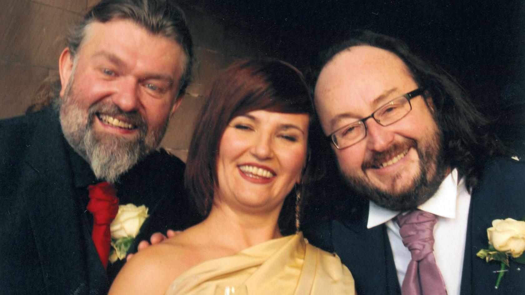 A picture taken at the wedding of Lili and Dave. Lili is in the middle of the shot and is wearing a cream wedding dress that comes off the shoulder. She is carrying a bouquet of orange and cream flowers and is carrying a glass of champagne in her hand. On her right is her husband, Dave. He is looking overjoyed and is smiling at the camera and wearing a purple tie, smart jacket and kilt. Si is on the left of the shot. He is wearing a red tie, smart jacket and a kilt and has a big smile on his face.