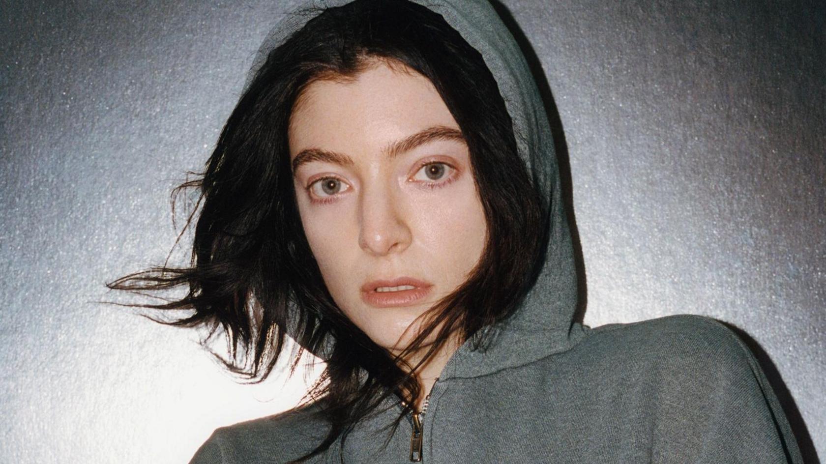 Lorde. She is pictured against a silver background wearing a grey hoodie with the hood up. She has long dark hair which is peeking out from underneath the hood.