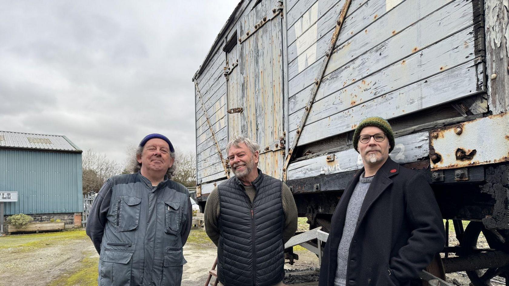 Three men standing in front of a blue-grey wooden railway wagon. They all have grey hair. On the left, the man wears a beanie and grey jacket. In the centre, the man is wearing a dark gilet, and on the right, the man is wearing a green beanie and black glasses.